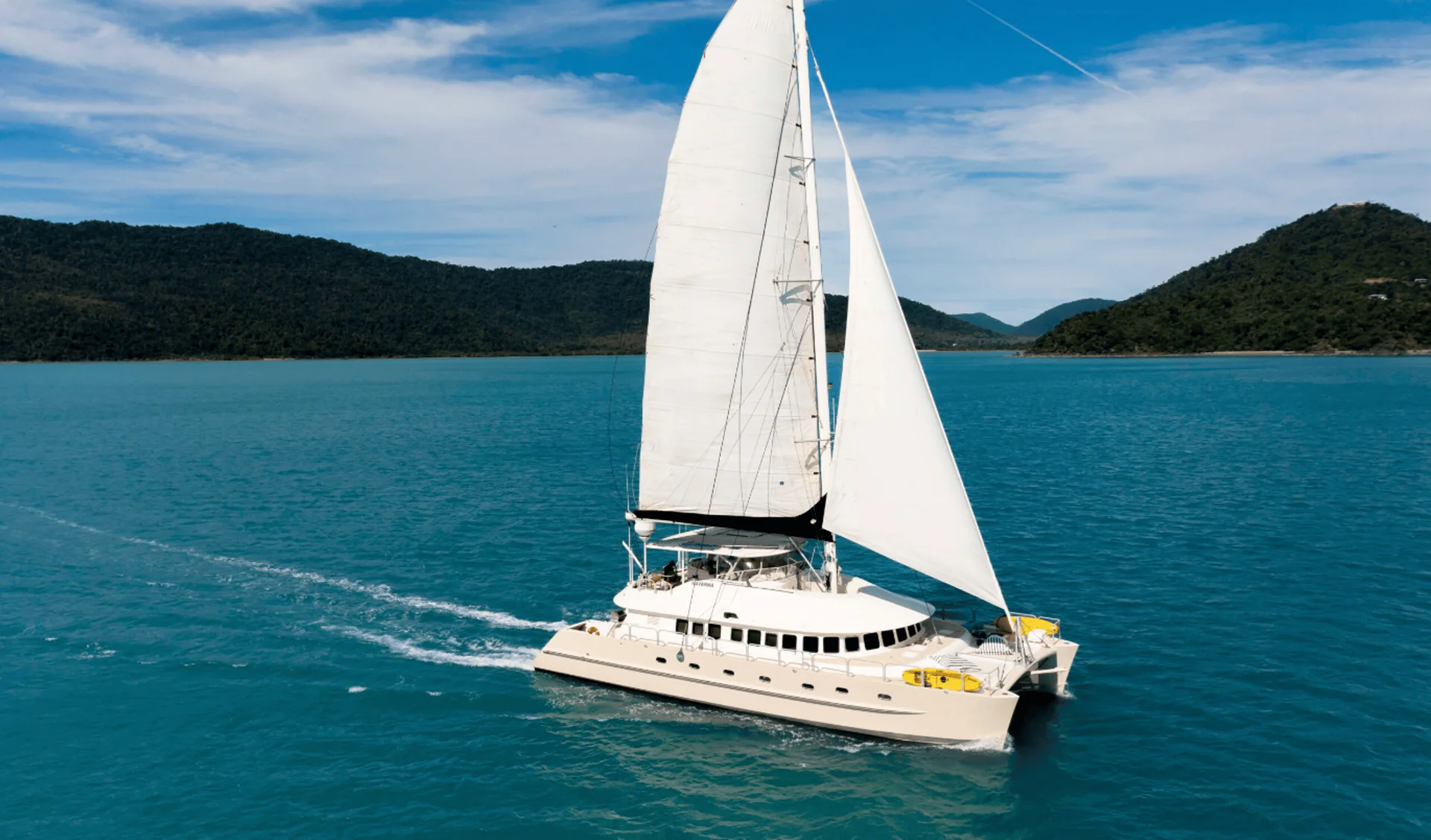 Havanna sailing across turquoise water off a forested island coastline.